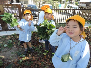写真：芋の葉っぱでネックレスをつけて微笑んでいる女児とはっぱをたくさん集めて手に持つ男児