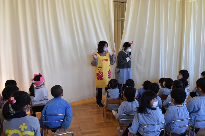 幕の前で話をする先生と先生の方を見つめる園児