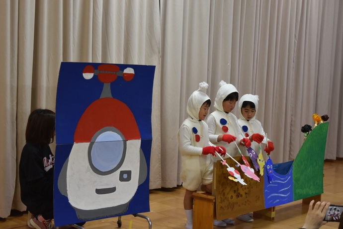 宇宙船の絵の隣で、白い衣装を着てペープサートをする園児たち