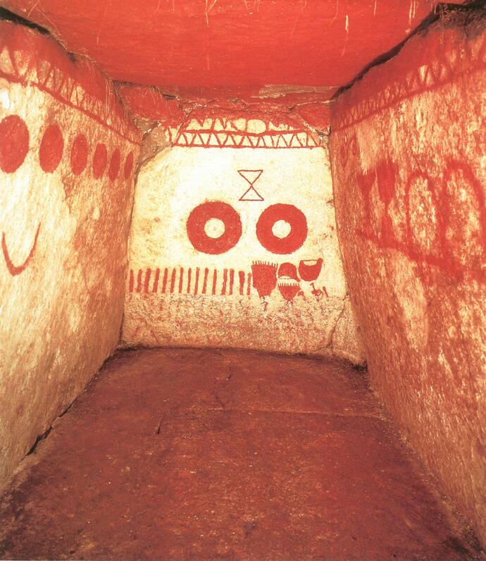 虎塚古墳石室内を入口側から撮影した写真。白い壁に赤い装飾が施されている。正面には、虎塚古墳の象徴的な図形、三角形2つを頂点が1つずつ接した図形の下に、ドーナツ型の赤い丸が2つ並んで描かれている。天井は全て赤く、左右の壁には正面とは違う様々な図形が描かれている。